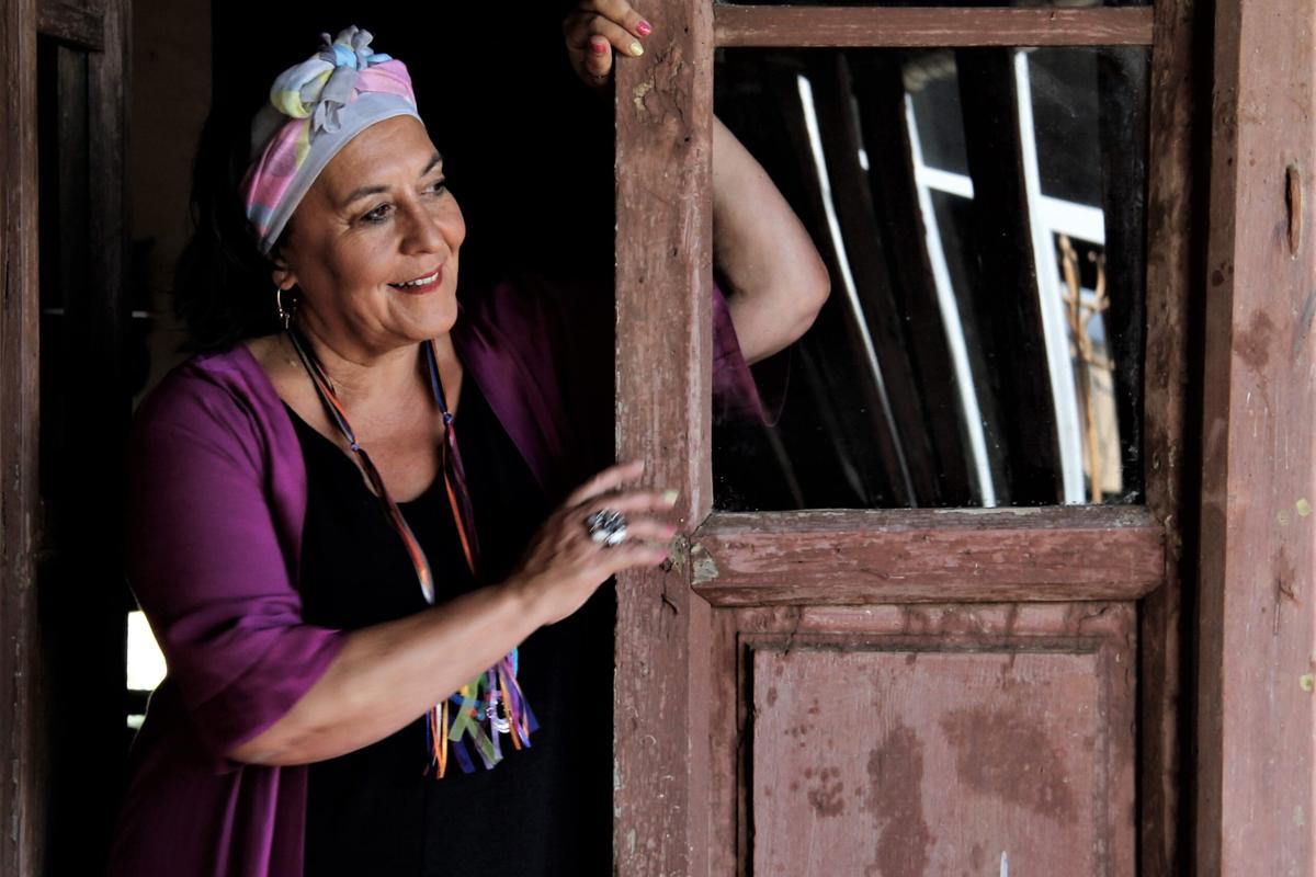 À esquerda de uma porta, já gasta pelos anos, está, de pé, uma mulher sorridente. Veste-se de negro e com um casaco roxo. Na cabeça, tem uma espécie de lenço, de cores claras e com um nó, ao centro.