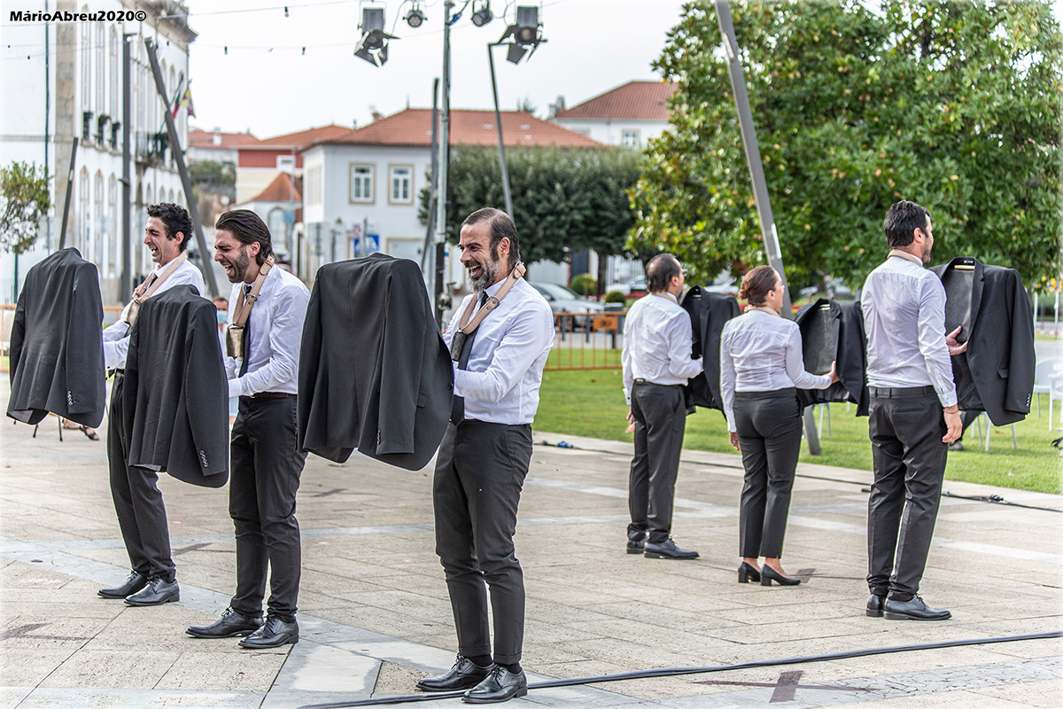 Ao ar livre, talvez numa praça, estão seis pessoas, formando duas linhas de três, de costas voltadas. Todas vestem calças escuras e camisa branca. Seguram, à sua frente, pela altura dos ombros, os seus blazers escuros - que estão pendurados em cabides.