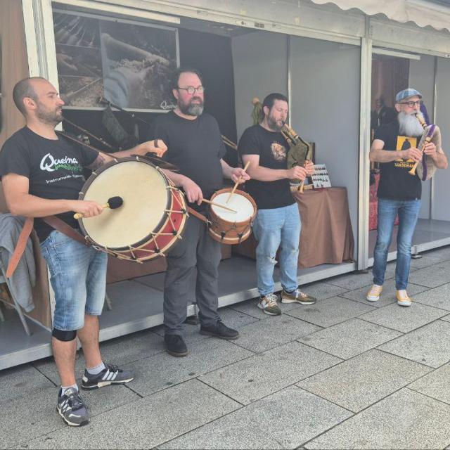 Quatro músicos estão, de pé, no que parece ser uma feira. Estão vestidos com t-shirt’s pretas e calças de ganga, três azuis e umas pretas. Dois deles tocam gaitas de foles e os demais tocam um bombo e um tambor.