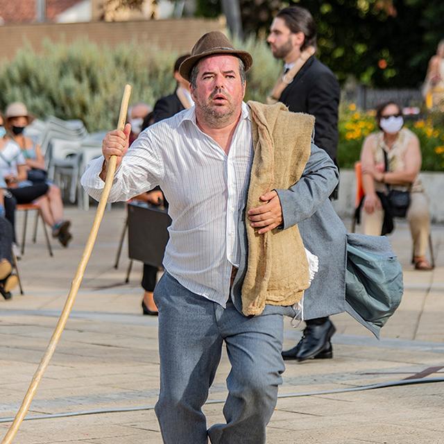 Ao ar livre, talvez numa praça, está, em primeiro plano, um homem de meia idade, que veste trajes tipicamente rurais, uma camisa branca, umas calças cinzentas, um chapéu. Tem também um casaco cinzento, vestindo-o apenas pela manga e pousado sobre o seu ombro direito, por cima do casaco, uma espécie de saco de sarapilheira.