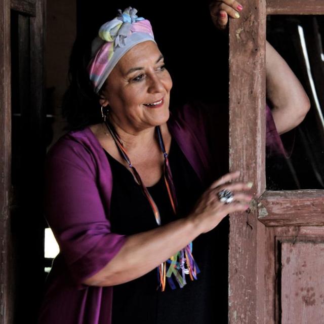 À esquerda de uma porta, já gasta pelos anos, está, de pé, uma mulher sorridente. Veste-se de negro e com um casaco roxo. Na cabeça, tem uma espécie de lenço, de cores claras e com um nó, ao centro.